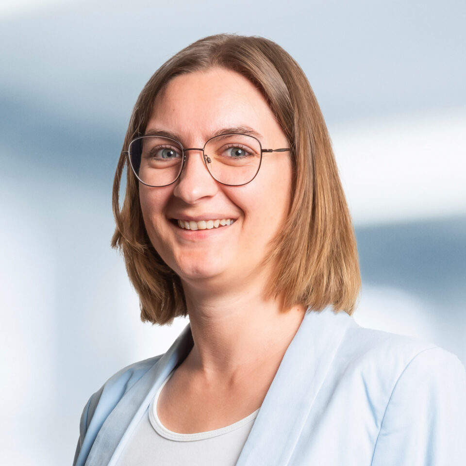 Das Bild zeigt ein professionelles Porträt einer Frau, die leicht zur Seite gedreht in die Kamera lächelt. Sie trägt eine runde, filigrane Brille und hat schulterlanges, glattes Haar, das sauber gescheitelt ist. Ihr Outfit wirkt hell und freundlich: ein hellblauer Blazer über einem weißen Oberteil. Der Hintergrund ist weich verlaufend in hellen Blau- und Weißtönen gehalten.