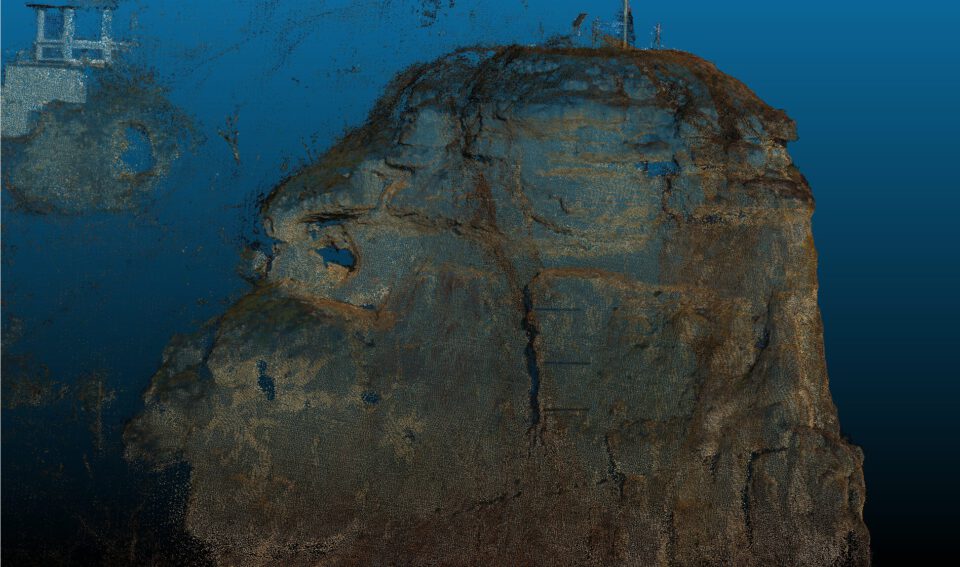Das Bild zeigt ein dreidimensionales Punktwolken-Modell einer steilen Felsformation vor einem gleichmäßig blauen Hintergrund. Der Fels ist massiv, unregelmäßig geformt und stark zerklüftet, mit deutlich erkennbaren Rissen, Schichtungen und Ausbrüchen in der Oberfläche.