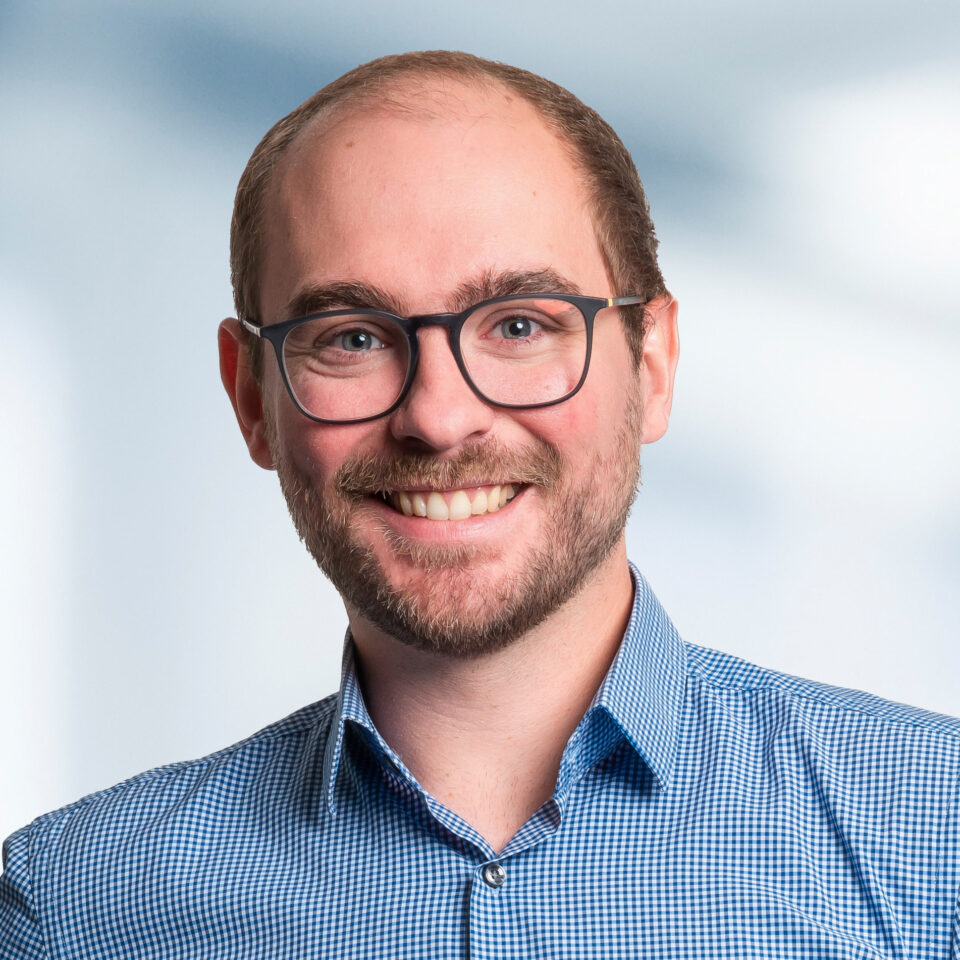 Das Bild zeigt ein professionelles Porträt eines Mannes, der direkt in die Kamera lächelt. Er trägt eine Brille mit dunklem Rahmen, hat einen kurzen Bart und eine leicht zurückgehende Haarlinie. Sein Hemd ist blau-weiß kariert.