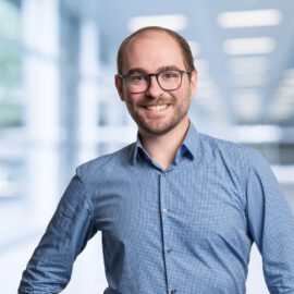 Das Bild zeigt eine Person in einem blau karierten Hemd mit langen Ärmeln, die leicht seitlich steht und eine Hand in die Hüfte gestützt hat. Der Hintergrund ist unscharf und zeigt eine helle, moderne Innenraumumgebung mit großen Fenstern und viel Licht.