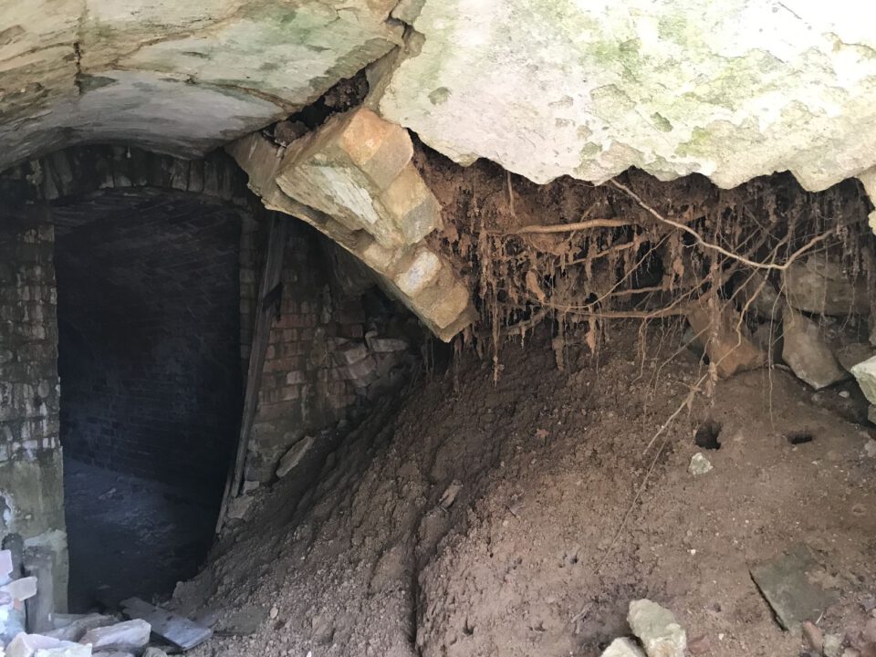 Das Bild zeigt einen teilweise eingestürzten Kellerbereich mit einer beschädigten Decke aus Stein und Mauerwerk. Aus der aufgebrochenen Decke hängen zahlreiche Wurzeln herab, und darunter hat sich ein großer Erdhaufen angesammelt. Im Hintergrund ist ein dunkler Durchgang aus Backsteinmauerwerk zu erkennen, der in einen weiteren Raum führt.