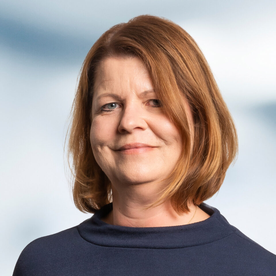 Das Bild zeigt ein professionelles Porträt einer Frau mit schulterlangem, leicht gewelltem, rötlich-braunem Haar. Sie blickt direkt in die Kamera und lächelt dezent, was einen ruhigen und freundlichen Eindruck vermittelt. Sie trägt ein dunkelblaues Oberteil mit einem weich fallenden, leicht erhöhten Ausschnitt. Der Hintergrund ist hell und neutral.