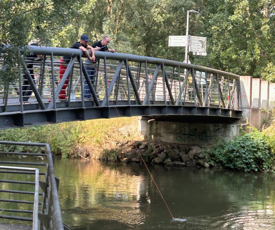 Das Bild zeigt eine Metallbrücke über einem kleinen Fluss, auf der mehrere Personen stehen und mit einer Leine oder einem Seil eine Probe aus dem Wasser entnehmen. Unter der Brücke fließt klares Wasser, umgeben von grüner Vegetation und Bäumen im Hintergrund. Die Szene wirkt wie eine praktische Messung oder Untersuchung in einer natürlichen Umgebung.