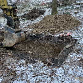 Das Bild zeigt eine Baustelle im Wald, auf der ein Bagger mit einer großen Schaufel ein rechteckiges Loch im Boden aushebt. Rund um die Grube liegt lockere Erde, und im Hintergrund sind weitere Erdhaufen sowie Bäume zu sehen. Der Boden ist leicht mit Schnee bedeckt, was auf kalte Witterung hinweist.