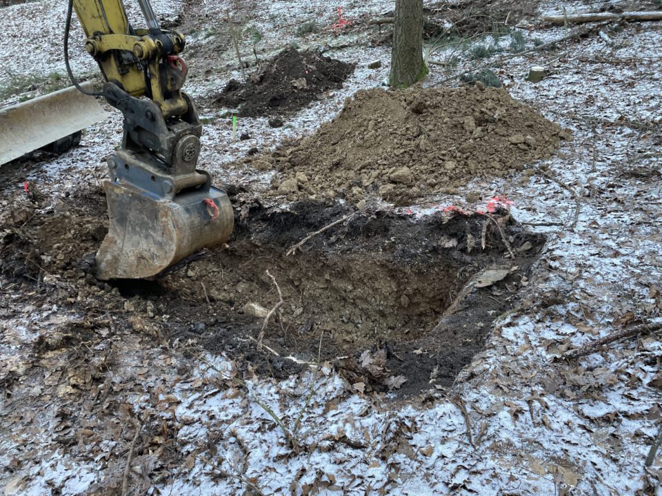 Das Bild zeigt eine Baustelle im Wald, auf der ein Bagger mit einer großen Schaufel ein rechteckiges Loch im Boden aushebt. Rund um die Grube liegt lockere Erde, und im Hintergrund sind weitere Erdhaufen sowie Bäume zu sehen. Der Boden ist leicht mit Schnee bedeckt, was auf kalte Witterung hinweist.