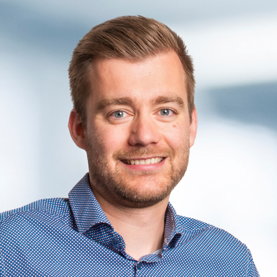 Das Bild zeigt ein freundlich lächelndes Porträt einer Person mit kurzem, seitlich gekämmtem Haar und leichtem Bart. Die Person trägt ein gemustertes Hemd in Blau und Weiß vor einem hellen, neutralen Hintergrund.