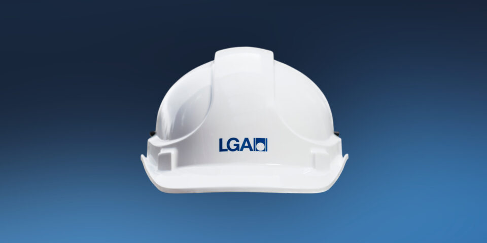 Das Bild zeigt einen weißen Schutzhelm vor einem dunkelblauen Hintergrund. Auf dem Helm ist das Logo der LGA.