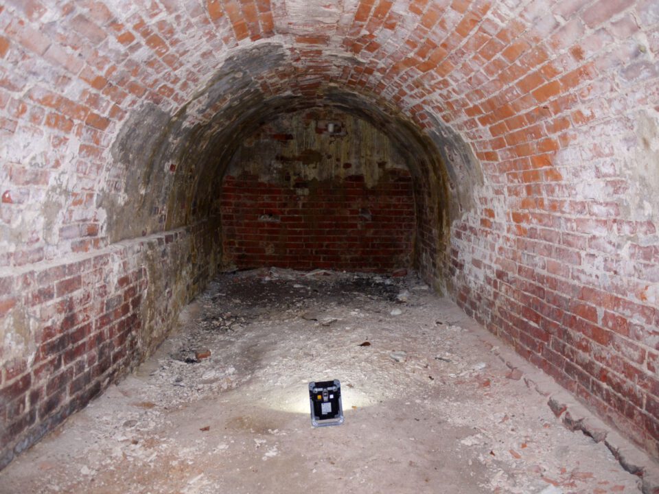 Das Bild zeigt einen kleinen, gemauerten Kellerraum mit Tonnengewölbe aus roten Ziegelsteinen. Die Wände und die Decke sind stark verschmutzt, verfärbt und stellenweise ausgebrochen. Am hinteren Ende ist eine zugemauerte Stirnwand aus Ziegeln zu erkennen.