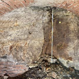 Das Bild zeigt eine gemauerte Kellerwand mit einem Gewölbebogen aus roten Ziegelsteinen. In der Mitte ist ein großer Bereich des natürlichen Felsgesteins sichtbar, das Risse und unterschiedliche Farbstrukturen aufweist. Am Boden liegen lose Steine und Erdmaterial, und eine Messlatte steht senkrecht zur Größenbestimmung.