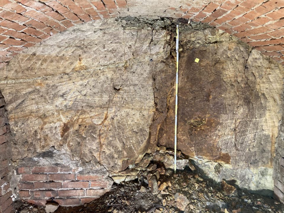 Das Bild zeigt eine gemauerte Kellerwand mit einem Gewölbebogen aus roten Ziegelsteinen. In der Mitte ist ein großer Bereich des natürlichen Felsgesteins sichtbar, das Risse und unterschiedliche Farbstrukturen aufweist. Am Boden liegen lose Steine und Erdmaterial, und eine Messlatte steht senkrecht zur Größenbestimmung.