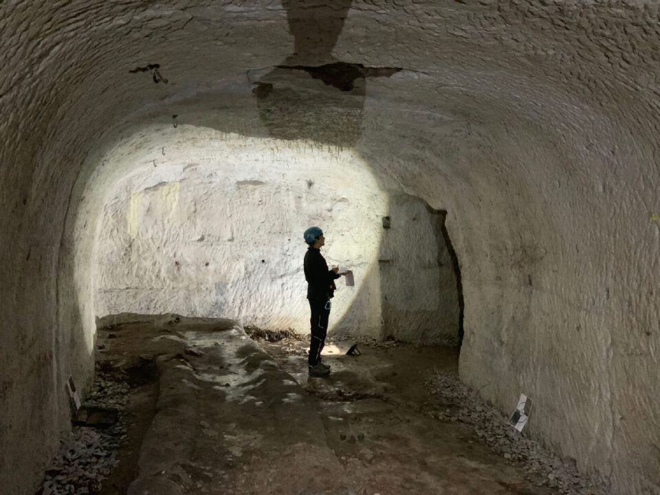Das Bild zeigt einen unterirdischen Raum mit hellen, grob behauenen Steinwänden und einer gewölbten Decke. Auf dem Boden liegen lose Steine und Erdmaterial, während eine Person mit Schutzhelm und Ausrüstung in der Mitte steht und die Wand untersucht. Der Raum wird von einer starken Lichtquelle beleuchtet, die die Struktur und Textur der Wände deutlich sichtbar macht.