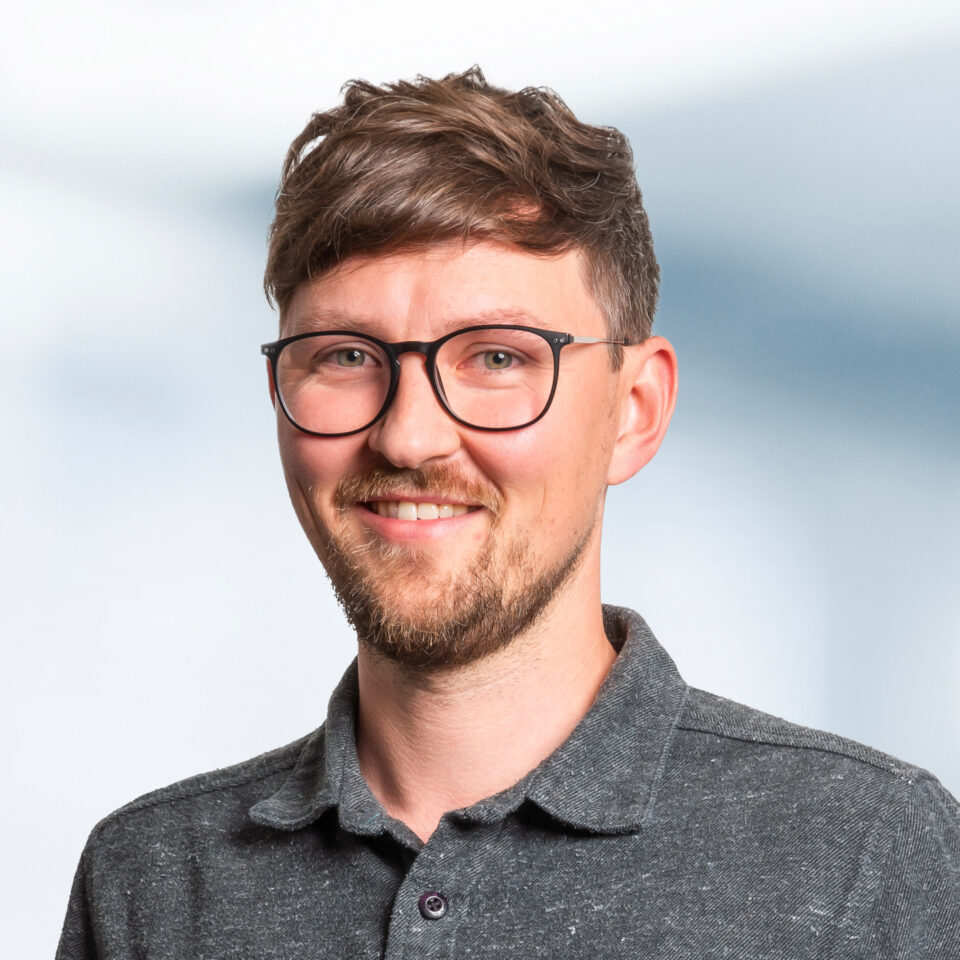 Das Bild zeigt ein freundlich lächelndes Porträt einer Person mit kurzem, gewelltem Haar, runder Brille und gepflegtem Bart. Die Person trägt ein dunkles Poloshirt und steht vor einem hellen, weich verlaufenden Hintergrund.
