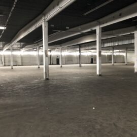 Das Bild zeigt eine große, leere Industriehalle mit einem weitläufigen Betonboden und zahlreichen weißen Stützpfeilern, die das Dach tragen. Die Halle ist gut beleuchtet durch eine Reihe von Lampen entlang der Decke, und im Hintergrund sind mehrere große Tore und Türen zu erkennen. Die Fläche wirkt sauber und frei von Maschinen oder Möbeln, was auf eine ungenutzte oder vorbereitete Lagerfläche hinweist.