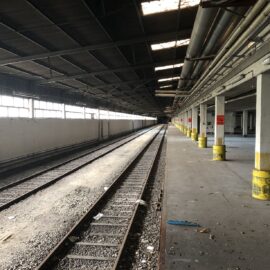 Das Bild zeigt eine große, überdachte Halle mit zwei parallel verlaufenden Eisenbahnschienen, die sich in die Tiefe des Raumes erstrecken. Links befindet sich eine lange weiße Wand mit Fenstern, während rechts eine Reihe von Säulen den offenen Bereich stützt, der teilweise mit gelben Markierungen versehen ist. Die Halle wirkt verlassen, der Boden ist stellenweise verschmutzt, und durch die Dachöffnungen fällt etwas Licht in den ansonsten dunklen Raum.