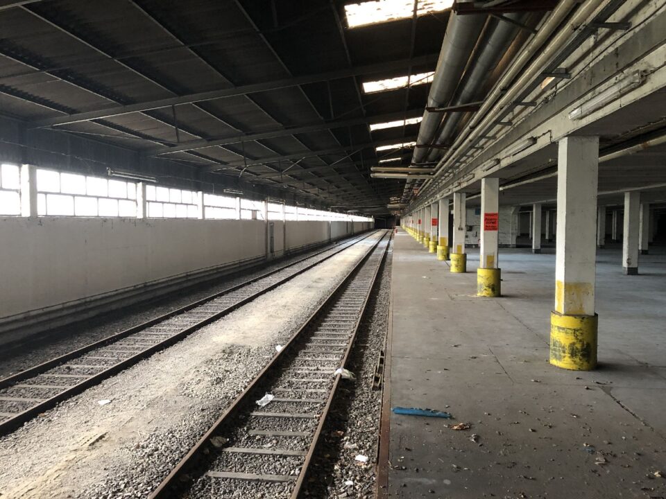 Das Bild zeigt eine große, überdachte Halle mit zwei parallel verlaufenden Eisenbahnschienen, die sich in die Tiefe des Raumes erstrecken. Links befindet sich eine lange weiße Wand mit Fenstern, während rechts eine Reihe von Säulen den offenen Bereich stützt, der teilweise mit gelben Markierungen versehen ist. Die Halle wirkt verlassen, der Boden ist stellenweise verschmutzt, und durch die Dachöffnungen fällt etwas Licht in den ansonsten dunklen Raum.