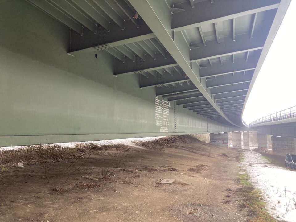 Das Bild zeigt die Unterseite einer großen Stahlbrücke mit massiven Trägern und Verstrebungen. Darunter verläuft eine breite, unbefestigte Fläche mit etwas Vegetation am Rand, die zu einem Fluss oder Wasserlauf führt, der rechts im Bild sichtbar ist. Im Hintergrund sind mehrere Betonpfeiler zu erkennen, die die Brücke stützen, und die Szene wirkt insgesamt wie eine technische Infrastruktur in einer natürlichen Umgebung.