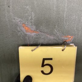 Das Bild zeigt eine Nahaufnahme einer grauen Metalloberfläche mit einer beschädigten Stelle, an der orangefarbene Grundierung oder Rost sichtbar ist. Vor der beschädigten Stelle befindet sich ein gelbes Schild mit der schwarzen Zahl 5, das an einem Ringbuch befestigt ist. Unten im Bild ist ein schwarzer Handschuh zu erkennen, der das Schild hält.