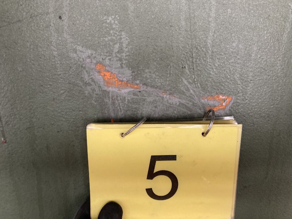 Das Bild zeigt eine Nahaufnahme einer grauen Metalloberfläche mit einer beschädigten Stelle, an der orangefarbene Grundierung oder Rost sichtbar ist. Vor der beschädigten Stelle befindet sich ein gelbes Schild mit der schwarzen Zahl 5, das an einem Ringbuch befestigt ist. Unten im Bild ist ein schwarzer Handschuh zu erkennen, der das Schild hält.
