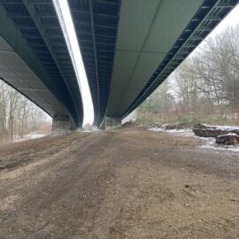 Das Bild zeigt eine breite, unbefestigte Fläche unter zwei parallel verlaufenden Brücken. Die Brücken sind aus Stahl und Beton gefertigt und werden von massiven Pfeilern getragen. Links und rechts der Fläche befinden sich Bäume und Sträucher, teilweise mit einer leichten Schneeschicht auf dem Boden, was auf winterliche Bedingungen hinweist. Auf der rechten Seite liegen mehrere große Baumstämme am Rand der Fläche.