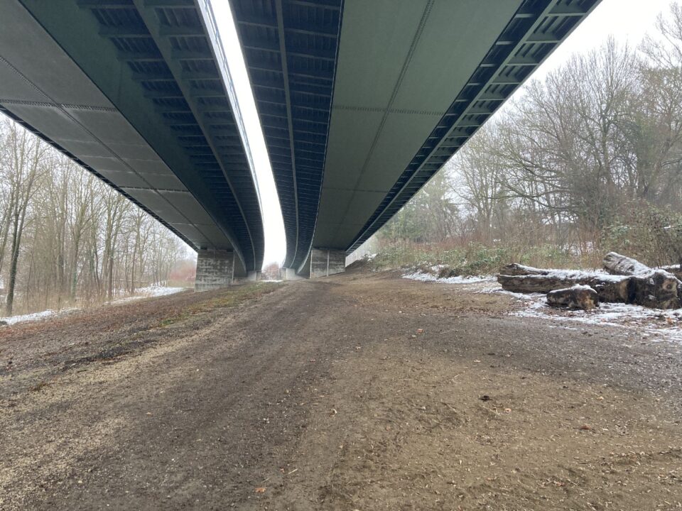Das Bild zeigt eine breite, unbefestigte Fläche unter zwei parallel verlaufenden Brücken. Die Brücken sind aus Stahl und Beton gefertigt und werden von massiven Pfeilern getragen. Links und rechts der Fläche befinden sich Bäume und Sträucher, teilweise mit einer leichten Schneeschicht auf dem Boden, was auf winterliche Bedingungen hinweist. Auf der rechten Seite liegen mehrere große Baumstämme am Rand der Fläche.