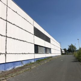 Das Bild zeigt die lange Seite eines großen, hellen Industriegebäudes mit einer glatten Fassade, die durch horizontale Linien strukturiert ist. Vor dem Gebäude verläuft eine asphaltierte Straße, deren Rand von vereinzelten Gräsern und kleinen Pflanzen bewachsen ist. Im Hintergrund sind Bäume, Laternen und ein klarer blauer Himmel zu sehen, was auf einen sonnigen Tag hinweist.