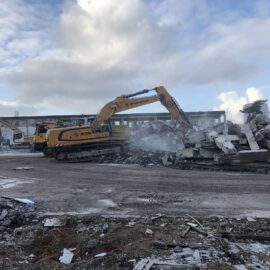 Auf dem Bild sind mehrere große gelbe Bagger zu sehen, die auf einer Baustelle arbeiten. Sie reißen Teile eines massiven Betongebäudes ab, wobei große Trümmer und Staubwolken entstehen. Im Vordergrund liegen verstreute Betonstücke und Schutt, während im Hintergrund die Reste der Gebäudestruktur sichtbar sind.