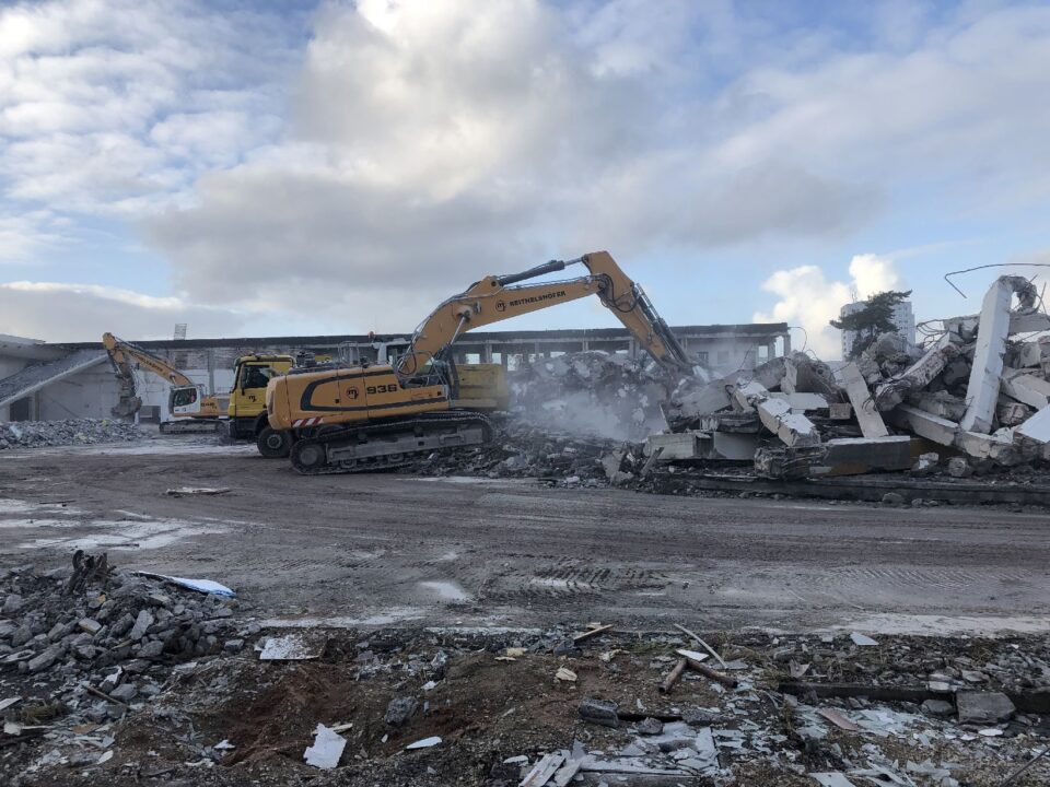 Auf dem Bild sind mehrere große gelbe Bagger zu sehen, die auf einer Baustelle arbeiten. Sie reißen Teile eines massiven Betongebäudes ab, wobei große Trümmer und Staubwolken entstehen. Im Vordergrund liegen verstreute Betonstücke und Schutt, während im Hintergrund die Reste der Gebäudestruktur sichtbar sind.