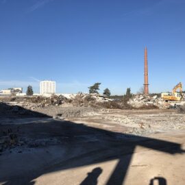 Das Bild zeigt eine große Abbruch- oder Baustelle mit weitgehend freigeräumter Fläche, auf der Betonreste und Schutt verteilt liegen. Im Hintergrund sind zwei gelbe Bagger zu sehen, die auf einem Schutthaufen arbeiten, sowie ein hoher, schlanker Schornstein, der aus der Szenerie herausragt. Dahinter befinden sich einige Bäume und ein weißes Hochhaus, während der Himmel klar und blau ist, was auf sonniges Wetter hinweist.