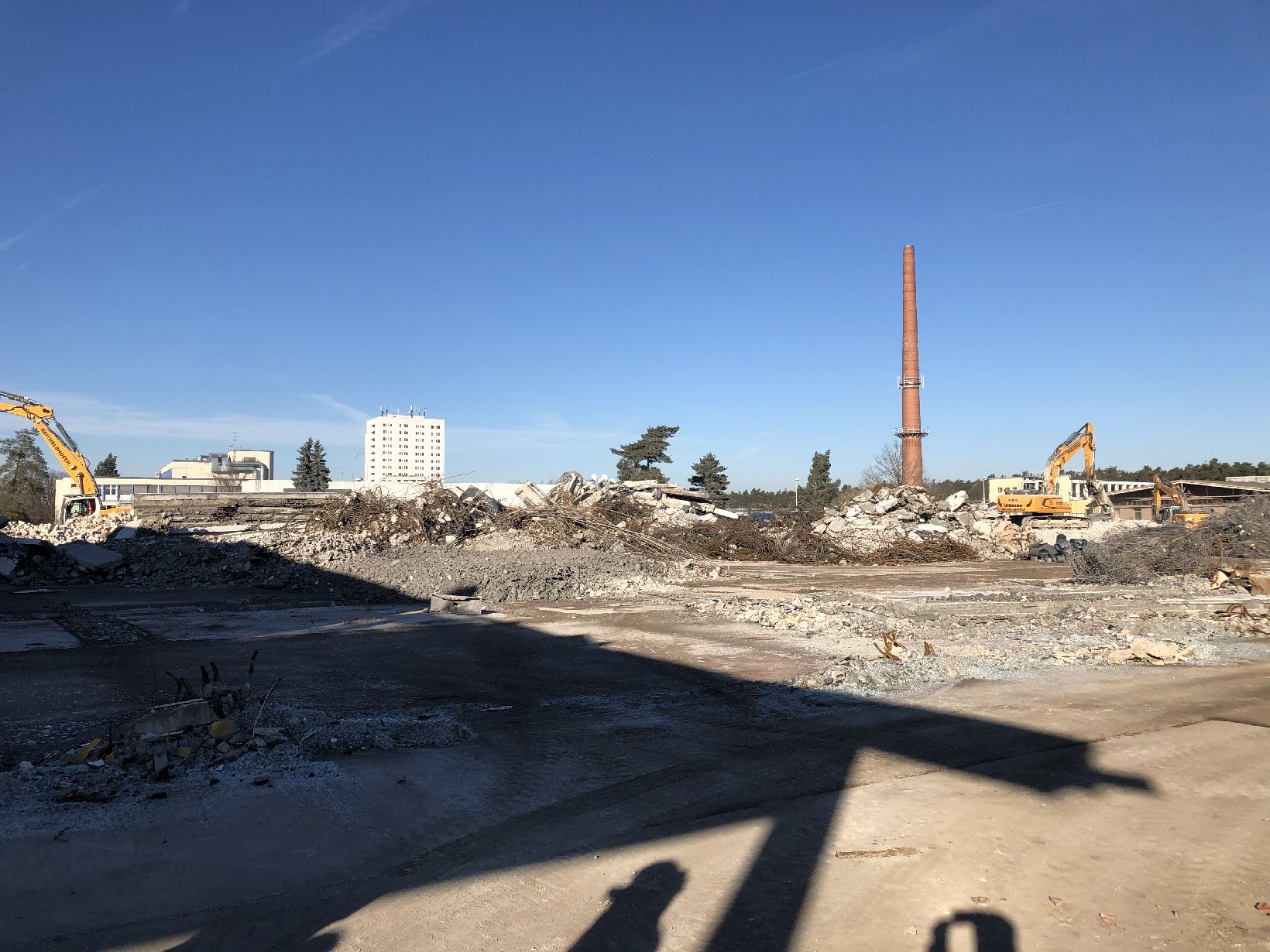Das Bild zeigt eine große Abbruch- oder Baustelle mit weitgehend freigeräumter Fläche, auf der Betonreste und Schutt verteilt liegen. Im Hintergrund sind zwei gelbe Bagger zu sehen, die auf einem Schutthaufen arbeiten, sowie ein hoher, schlanker Schornstein, der aus der Szenerie herausragt. Dahinter befinden sich einige Bäume und ein weißes Hochhaus, während der Himmel klar und blau ist, was auf sonniges Wetter hinweist.