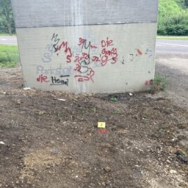 Das Bild zeigt den unteren Bereich eines großen Betonpfeilers einer Brücke, der mit bunten Graffiti in Rot, Schwarz und Blau besprüht ist. Davor befindet sich eine unbefestigte Fläche aus Erde und Kies mit vereinzelten kleinen Pflanzen. Auf dem Boden liegt ein gelbes Markierungsschild mit der Zahl 2 neben einem roten Punkt, was auf eine vermessene oder untersuchte Stelle hinweist. Im Hintergrund ist eine Straße sowie grüne Vegetation zu erkennen.