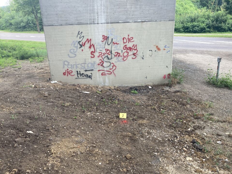 Das Bild zeigt den unteren Bereich eines großen Betonpfeilers einer Brücke, der mit bunten Graffiti in Rot, Schwarz und Blau besprüht ist. Davor befindet sich eine unbefestigte Fläche aus Erde und Kies mit vereinzelten kleinen Pflanzen. Auf dem Boden liegt ein gelbes Markierungsschild mit der Zahl 2 neben einem roten Punkt, was auf eine vermessene oder untersuchte Stelle hinweist. Im Hintergrund ist eine Straße sowie grüne Vegetation zu erkennen.