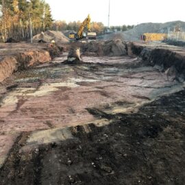 Das Bild zeigt eine große Baugrube mit freigelegten Erdschichten in unterschiedlichen Farben, darunter auffällige rötliche und gelbliche Bereiche. In der Mitte der Grube steht ein einzelner Betonfundamentrest, während im Hintergrund ein gelber Bagger auf einer erhöhten Fläche arbeitet. Rund um die Grube sind Erdhaufen aufgeschüttet, und rechts befinden sich Baucontainer sowie ein Zaun, der das Gelände abgrenzt. Links ist ein Waldstück mit hohen Bäumen zu sehen, was auf eine Baustelle in naturnaher Umgebung hinweist.