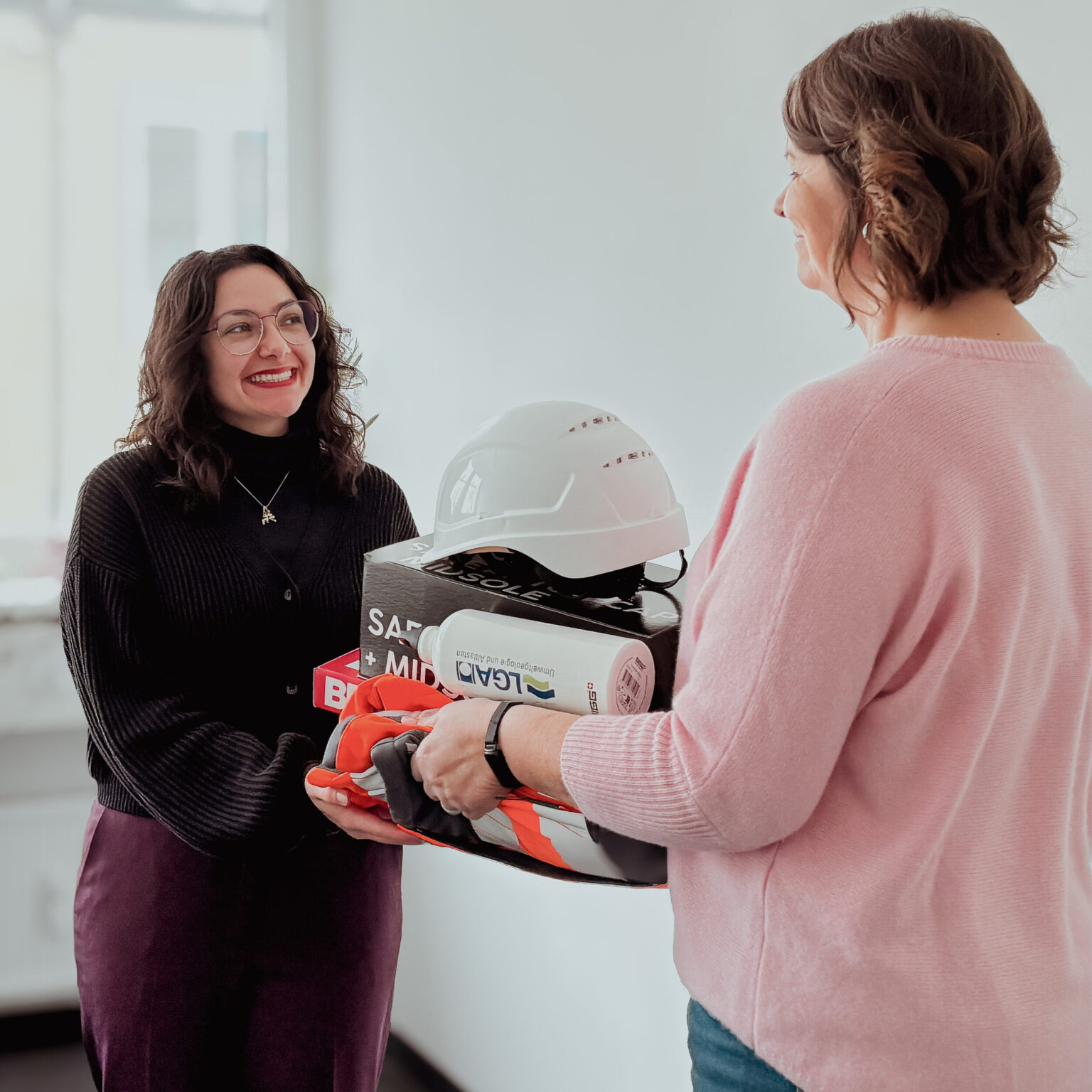 Zwei Personen stehen sich in einem hellen Innenraum gegenüber, während eine von ihnen ein Set aus Sicherheitsausrüstung überreicht. Auf dem Stapel liegen unter anderem ein Schutzhelm, Handschuhe und weitere Arbeitsmaterialien. Die Szene wirkt wie eine Übergabe oder Einweisung im Arbeitsumfeld.
