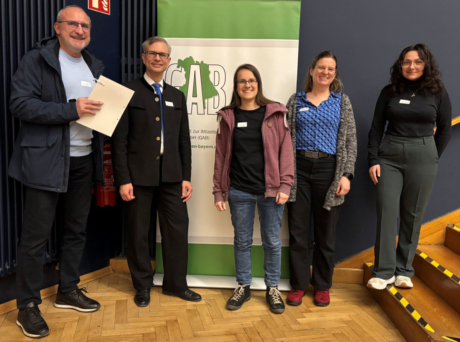 Eine Gruppe von fünf Personen steht in einem Innenraum vor einem grünen Banner der GAB. Die Personen posieren nebeneinander auf einem Parkettboden und tragen Namensschilder.