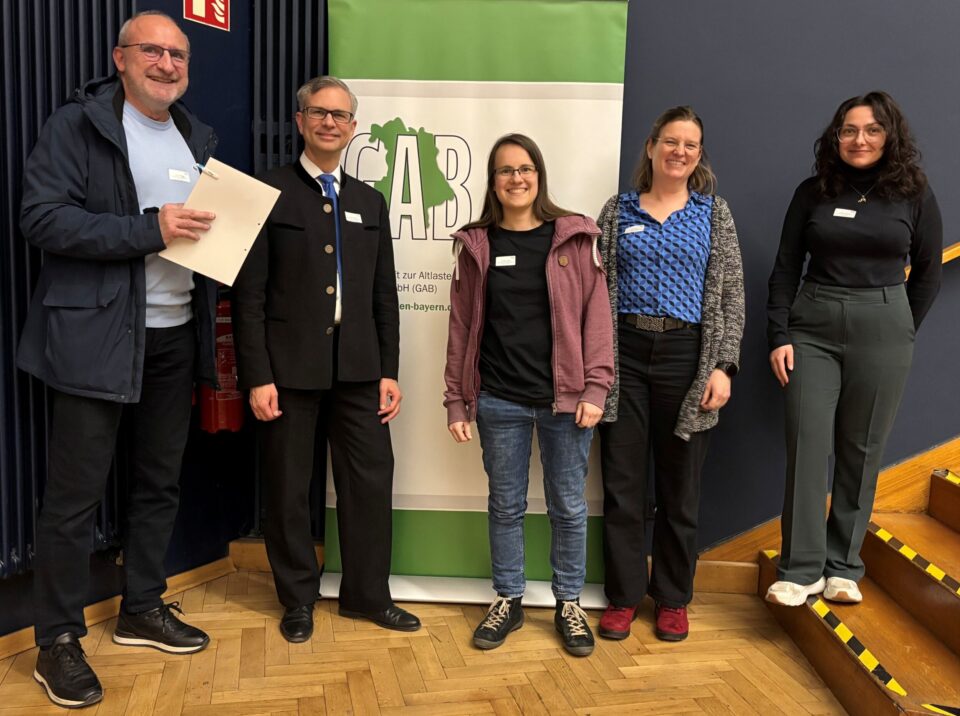 Eine Gruppe von fünf Personen steht in einem Innenraum vor einem grünen Banner der GAB. Die Personen posieren nebeneinander auf einem Parkettboden und tragen Namensschilder.