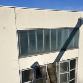 Das Bild zeigt die obere Hälfte eines hellen Gebäudes mit großen, rechteckigen Glasfenstern in der Mitte und darunter einer Reihe von Türen. Die Fassade ist schlicht und leicht verschmutzt, während auf der rechten Seite ein deutlicher Schatten eines großen Baugeräts auf die Wand fällt. Im Hintergrund sind Teile eines weiteren Gebäudes sowie ein klarer blauer Himmel zu erkennen.