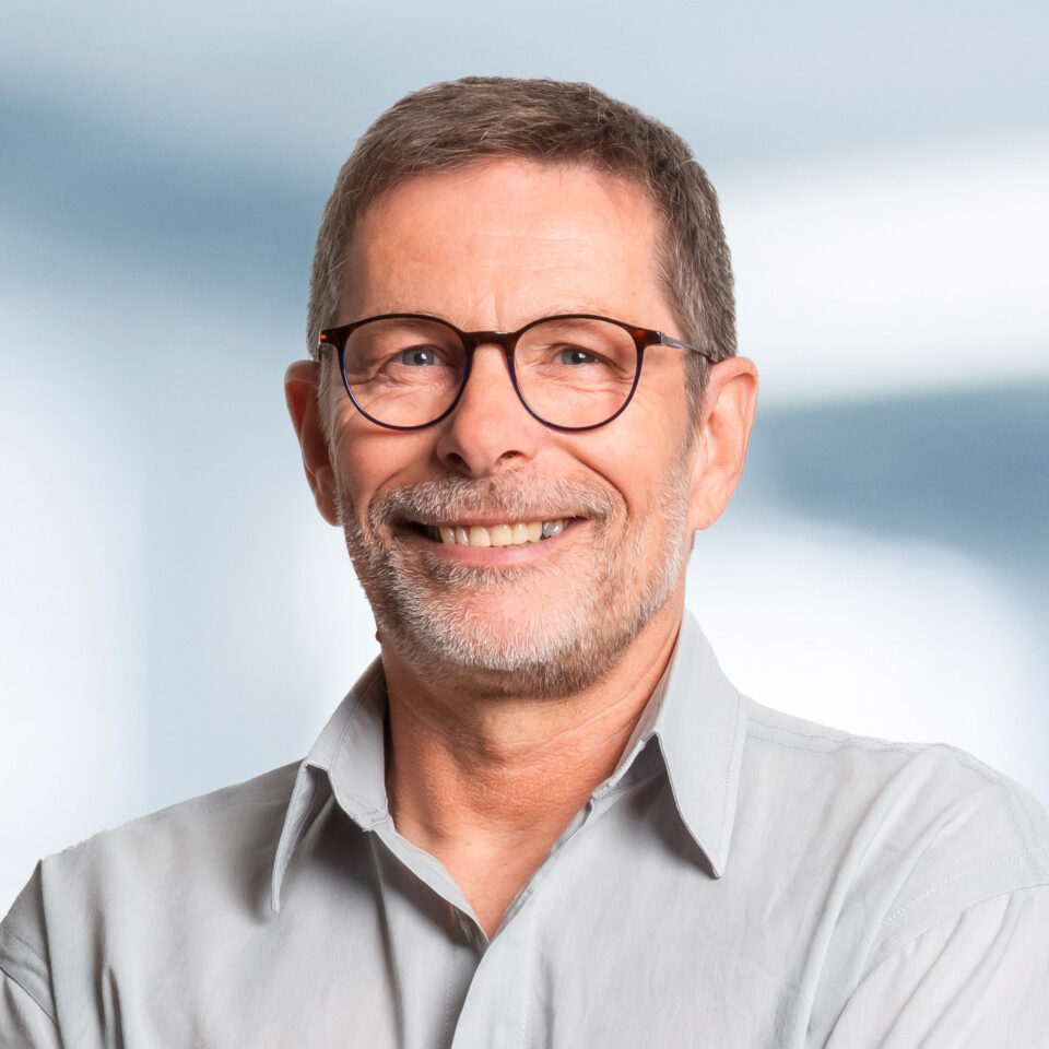 Das Bild zeigt ein freundlich lächelndes Porträt einer Person mit kurzem grauem Haar, Brille und gepflegtem Bart vor einem hellen, neutralen Hintergrund. Die Person trägt ein helles Hemd und wirkt offen und sympathisch.