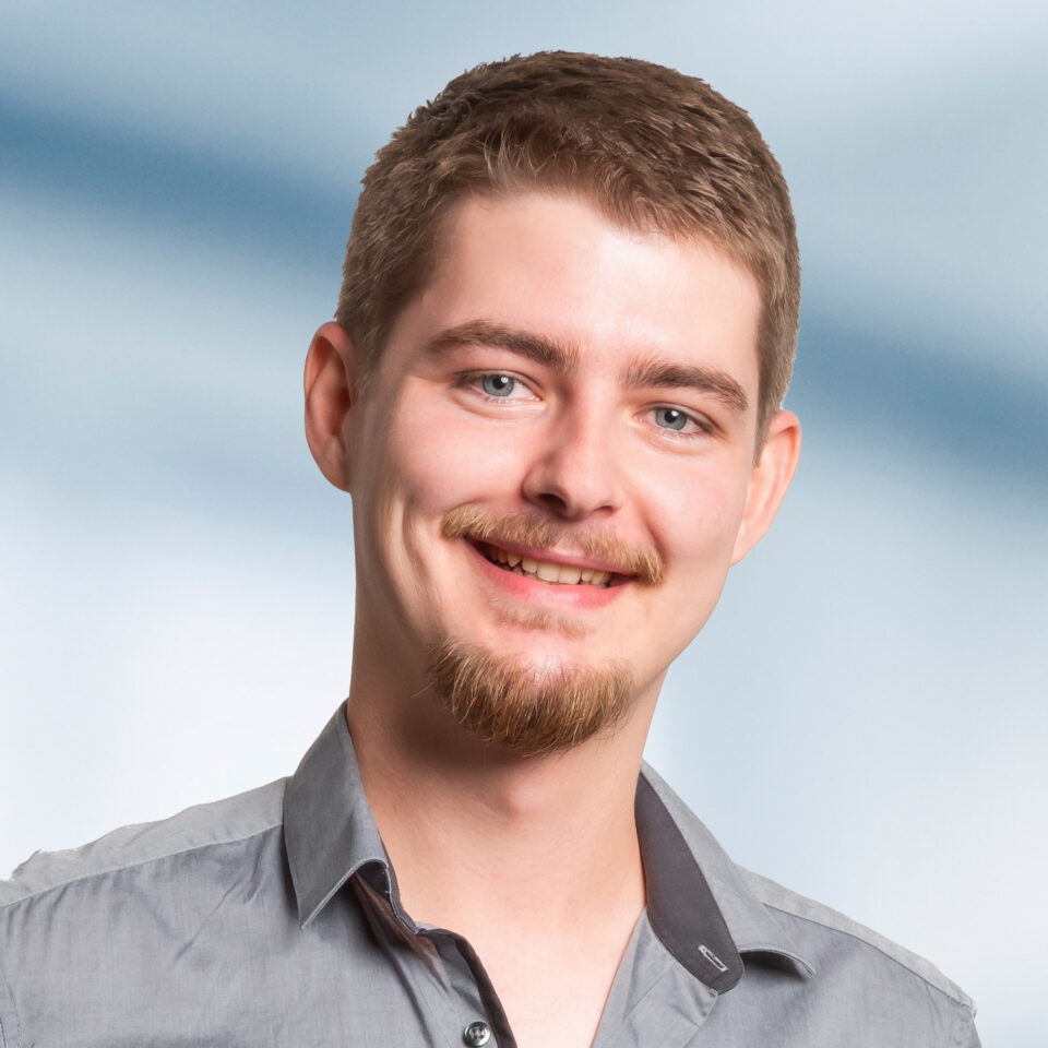 Das Bild zeigt ein freundlich lächelndes Porträt einer Person mit kurzem Haar, hellem Bart und Schnurrbart vor einem hellen, neutralen Hintergrund. Die Person trägt ein graues Hemd mit leicht geöffnetem Kragen.