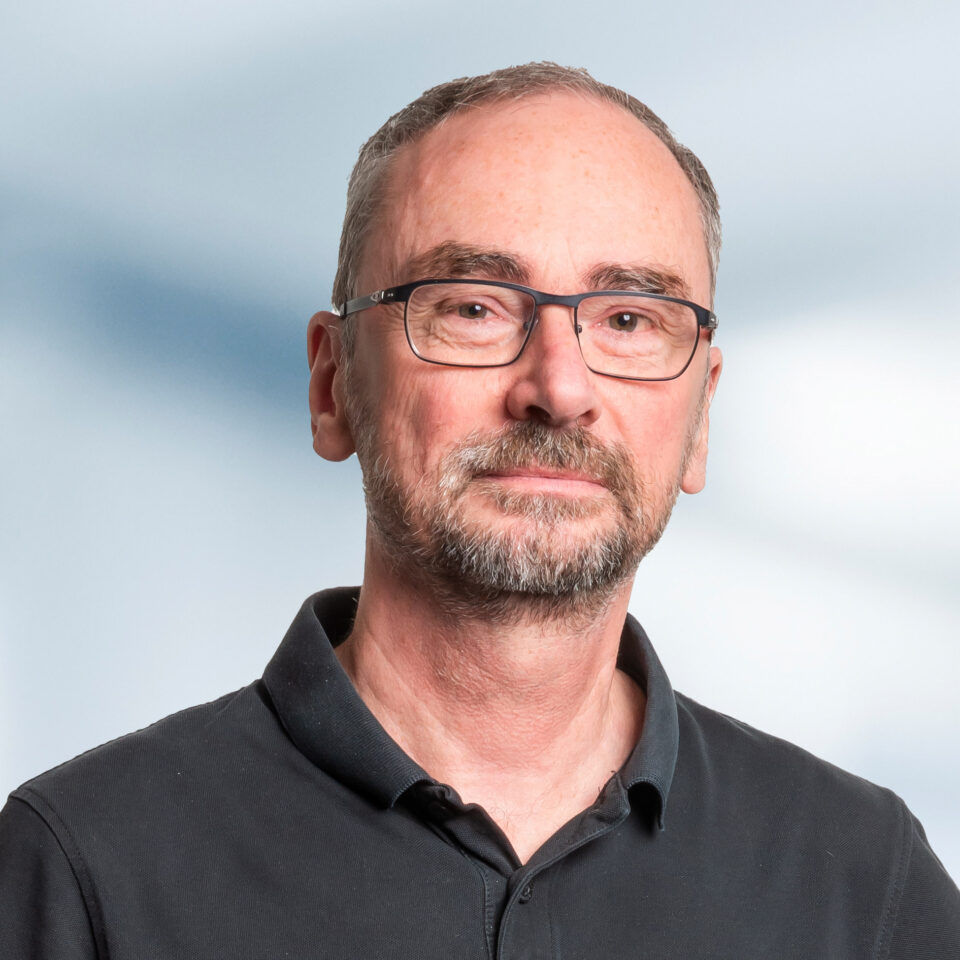 Das Bild zeigt ein sachliches Porträt einer Person mit kurzer Haarlänge, Brille und gepflegtem Bart vor hellem, neutralem Hintergrund. Die Person trägt ein dunkles Poloshirt.
