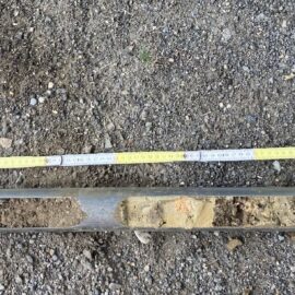 Das Bild zeigt eine Bodenprobe, die in einem länglichen Metallrohr entnommen wurde und auf einem kiesigen Untergrund liegt. Neben der Probe ist ein Maßstab mit Zentimeterangaben ausgelegt, um die Länge der entnommenen Schicht zu dokumentieren. Rechts oben befindet sich ein gelbes Schild mit der schwarzen Zahl 1, das die Probe eindeutig markiert.