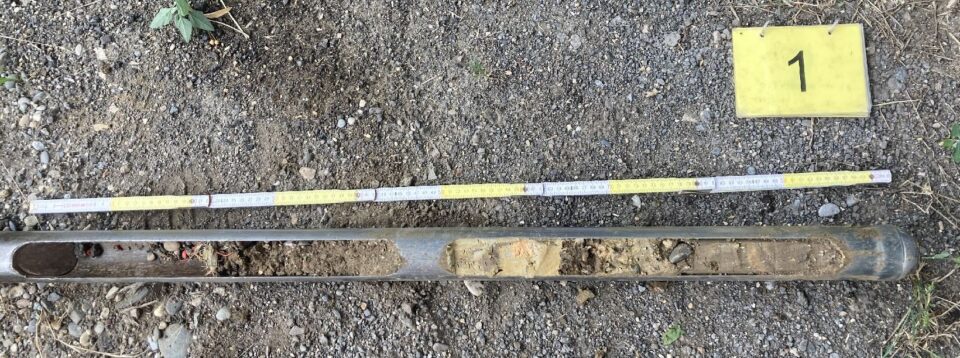 Das Bild zeigt eine Bodenprobe, die in einem länglichen Metallrohr entnommen wurde und auf einem kiesigen Untergrund liegt. Neben der Probe ist ein Maßstab mit Zentimeterangaben ausgelegt, um die Länge der entnommenen Schicht zu dokumentieren. Rechts oben befindet sich ein gelbes Schild mit der schwarzen Zahl 1, das die Probe eindeutig markiert.