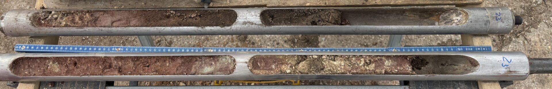 Auf dem Bild ist eine detaillierte Nahaufnahme von zwei längs aufgeschnittenen Bohrkernen zu sehen, die in metallischen Halterungen liegen. Die Bohrkerne zeigen unterschiedliche Boden- und Gesteinsschichten mit variierenden Farben, Körnungen und einzelnen Steinen, ergänzt durch einen darunterliegenden Maßstab zur Längenorientierung. Die Anordnung dient der genauen Dokumentation und Analyse der entnommenen Erdschichten.