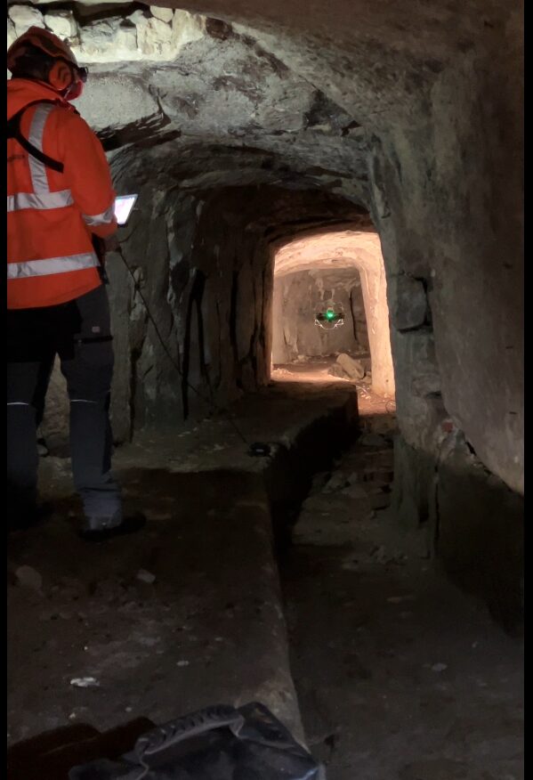 Das Bild zeigt einen schmalen, unterirdischen Gang mit grob behauenen Felswänden und unebenem Boden. Im Vordergrund steht eine Person in orangefarbener Schutzkleidung und Helm, die ein Steuergerät bedient. In der Mitte des Ganges schwebt eine kleine Drohne mit grün leuchtenden Positionslichtern, die für eine Inspektion eingesetzt wird.