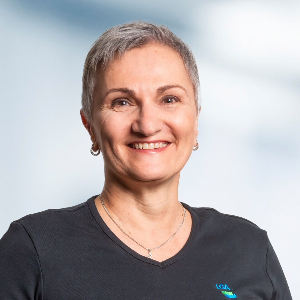 Das Bild zeigt ein freundlich lächelndes Porträt einer Person mit kurzem grauem Haar vor hellem, neutralem Hintergrund. Die Person trägt ein schwarzes T-Shirt, kleine Ohrringe und eine feine Kette.