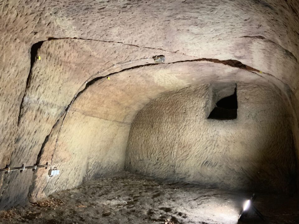 Das Bild zeigt einen unterirdischen Raum mit grob behauenen, hellen Steinwänden und einer gewölbten Decke. In der Decke sind mehrere Risse und Spalten sichtbar, die mit kleinen gelben Markierungen versehen wurden, vermutlich zur Dokumentation. Auf dem Boden liegt lose Erde, und eine starke Lichtquelle beleuchtet den Raum, wodurch die Struktur und die unregelmäßige Oberfläche der Wände deutlich hervortreten.