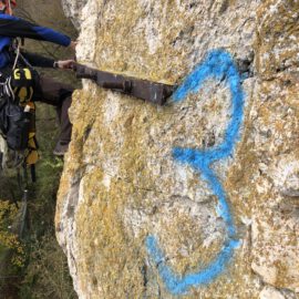 Das Bild zeigt eine Person, die mit Kletterausrüstung an einer steilen Felswand arbeitet. Die Felsoberfläche ist hell und mit gelblichen Flechten bedeckt, und darauf ist eine große blaue Markierung zu sehen. Die Person ist mit Seilen gesichert und hantiert an einem metallischen Bauteil, während im Hintergrund unscharf Bäume zu erkennen sind.