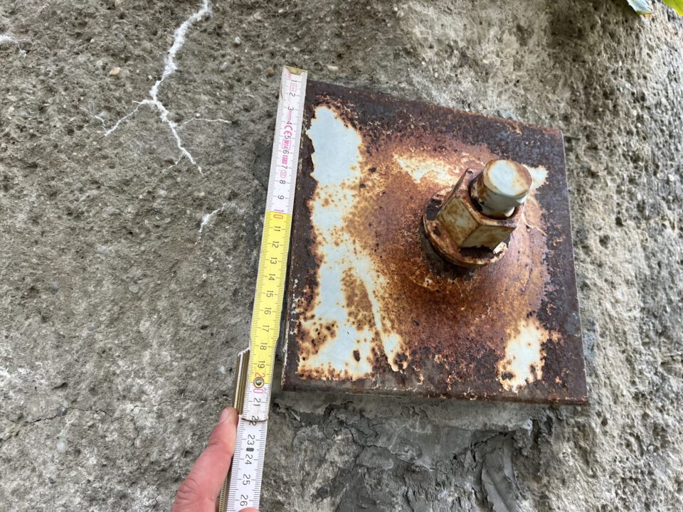 Das Bild zeigt eine stark korrodierte Metallplatte, die auf einer rauen Felsoberfläche befestigt ist. Neben der Platte wird ein Maßband angelegt, um die Größe zu überprüfen; die Skala ist deutlich sichtbar. Die Metalloberfläche weist Rostflecken und Abnutzungsspuren auf, was auf eine lange Witterungseinwirkung hinweist.