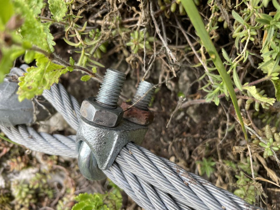 Das Bild zeigt eine Nahaufnahme einer Seilklemme an einem gespannten Stahlseil in natürlicher Umgebung. Im Zentrum ist eine grau verzinkte Seilklemme zu sehen, die das mehrdrähtige Stahlseil fixiert. Auf der Klemme befindet sich eine Metallmutter, die deutlich korrodiert und rostbraun verfärbt ist. Daneben ragt ein zweiter Gewindebolzen nach oben, ebenfalls mit Rostspuren.