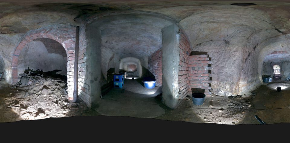 Das Bild zeigt ein Kugelpanorama eines unterirdischen Felsenkellers mit mehreren gewölbten Räumen. Die Wände bestehen aus grob behauenem Stein, teilweise mit gemauerten Bögen und Trennwänden aus Ziegeln. Auf dem Boden liegen verschiedene Eimer und Behälter, und die Beleuchtung hebt die Struktur der Wände sowie die unregelmäßige Oberfläche des Bodens deutlich hervor.