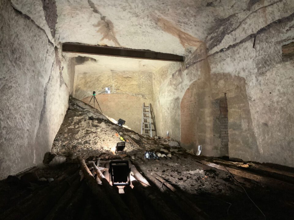 Das Bild zeigt einen großen unterirdischen Raum mit hellen, grob behauenen Wänden und einer hohen Decke. In der Mitte liegt ein großer Schuttkegel, auf dem ein Laserscanner auf einem Stativ positioniert ist, daneben eine Leiter, die an der Wand lehnt. Mehrere Lichtquellen beleuchten den Raum, und auf dem Boden sind Holzstämme sowie lose Materialien verteilt, was auf eine Vermessung oder Dokumentation der Struktur hinweist.